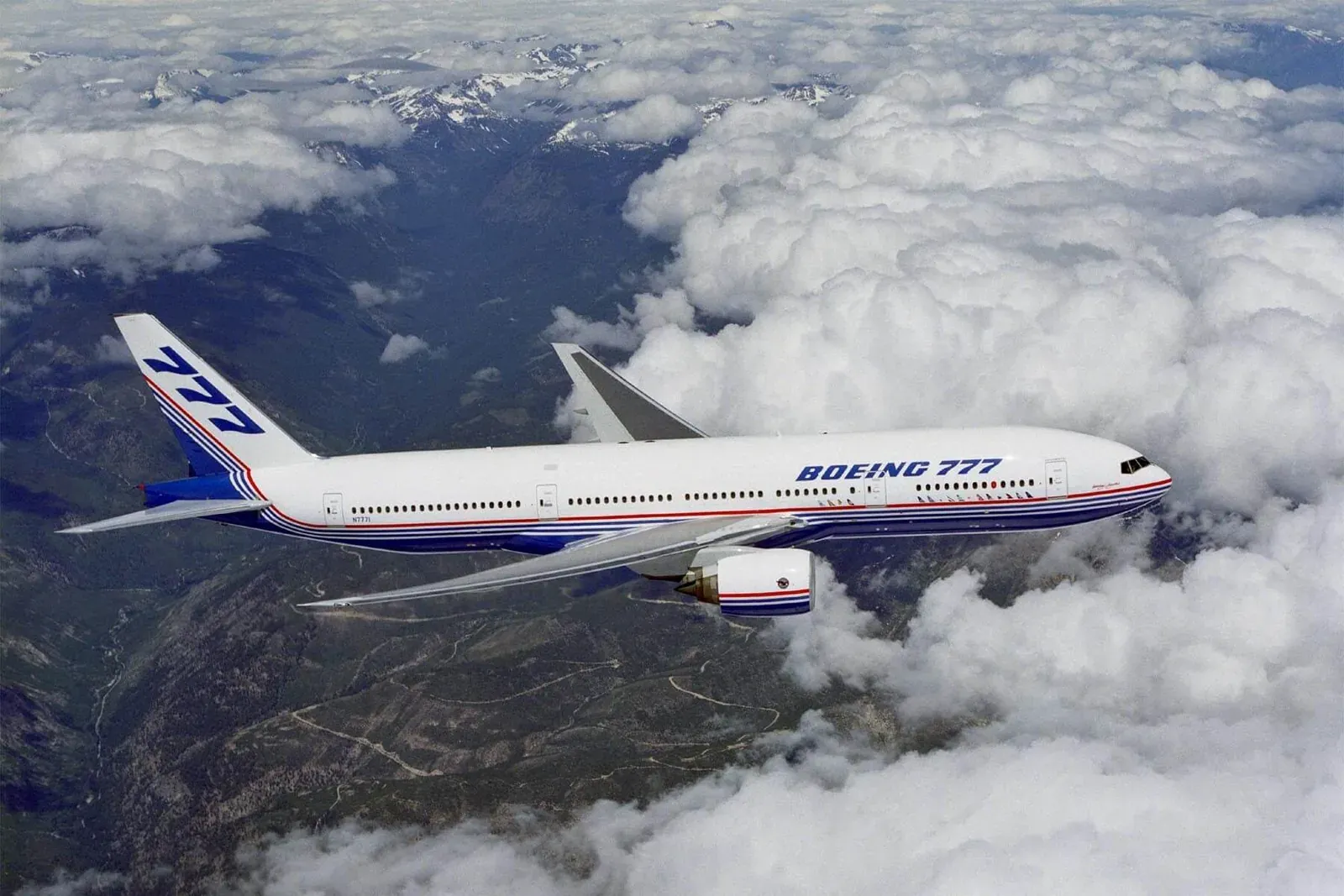A Boeing 777 in the original Boeing livery cruising above snow-capped mountains and cloud.