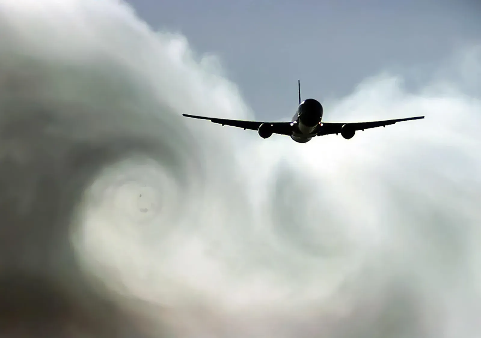 The rear view of an airliner climbing through swirling clouds, wingtip vortices spiralling behind it.