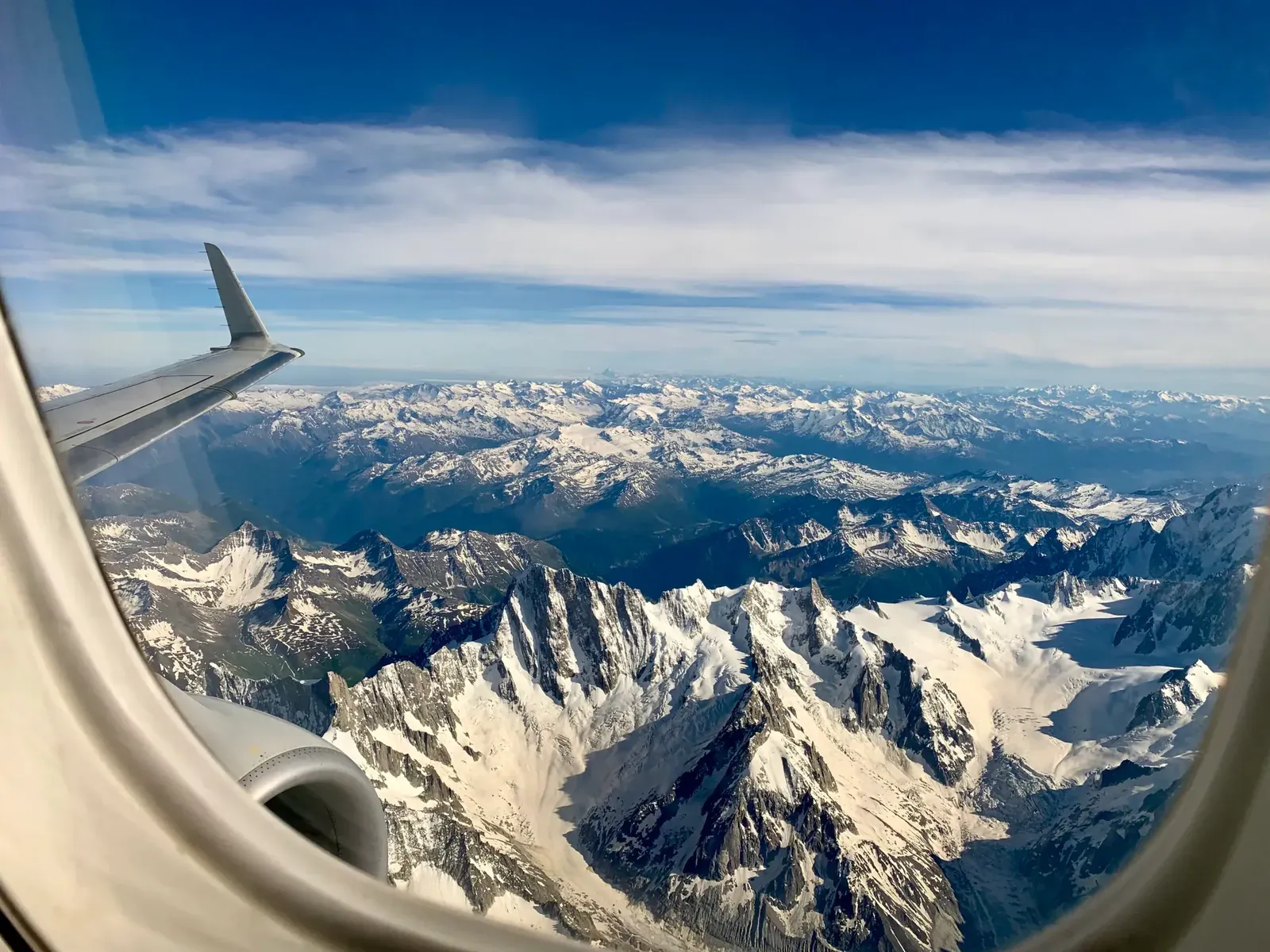 The view from an airplane window, its rounded corner framing a wing and snow-covered mountains far below.